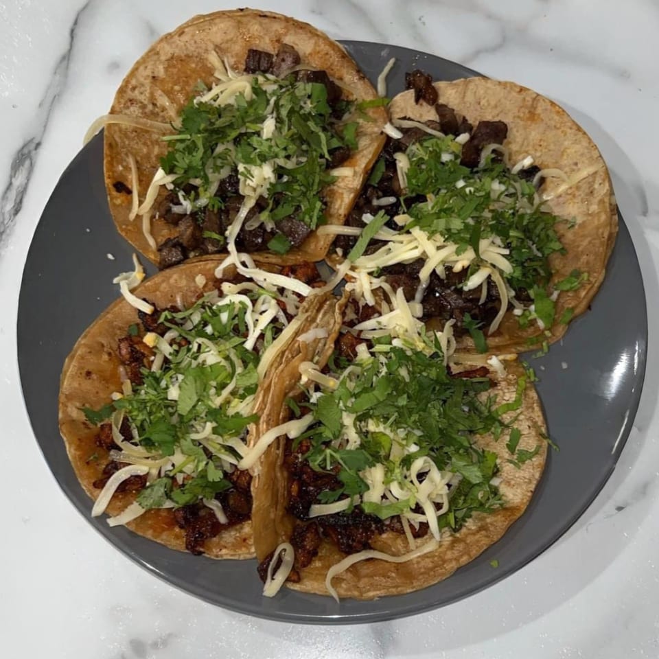 Four Mexican tacos with meat, shredded cheese, and cilantro on a gray plate.