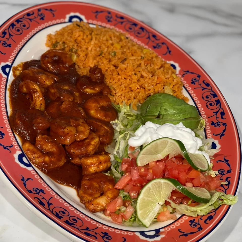Mexican shrimp dish with red sauce, rice, avocado, sour cream, and lime on a decorative plate.