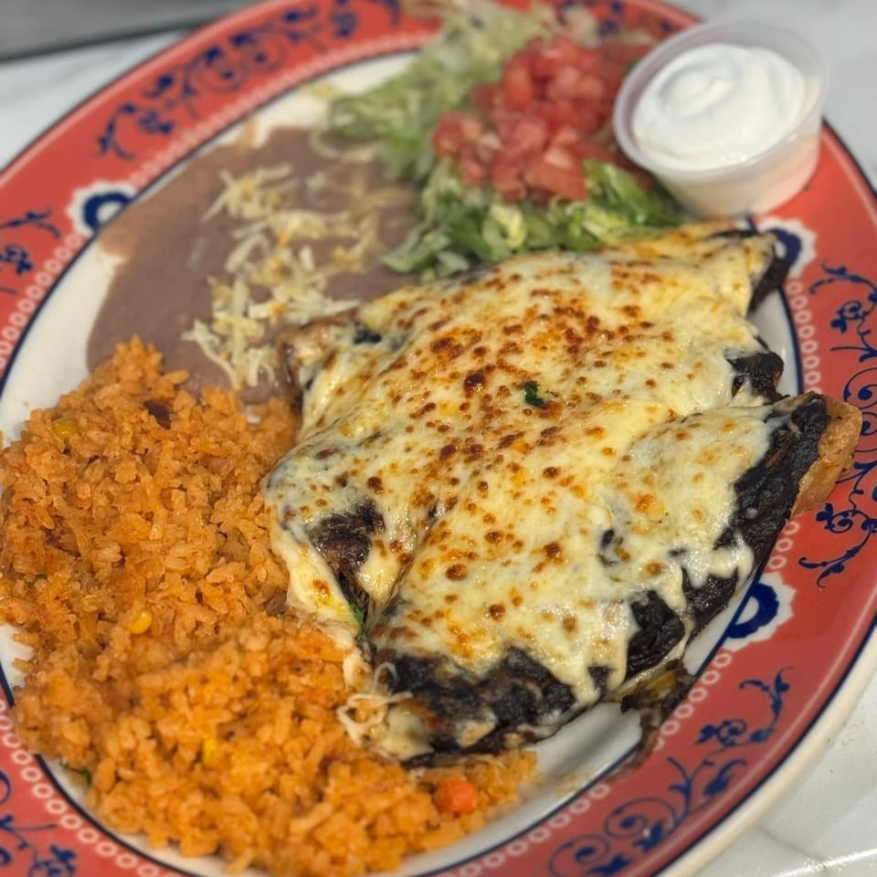 A Mexican dish with two cheesy enchiladas, rice, refried beans, lettuce, tomatoes, and sour cream.