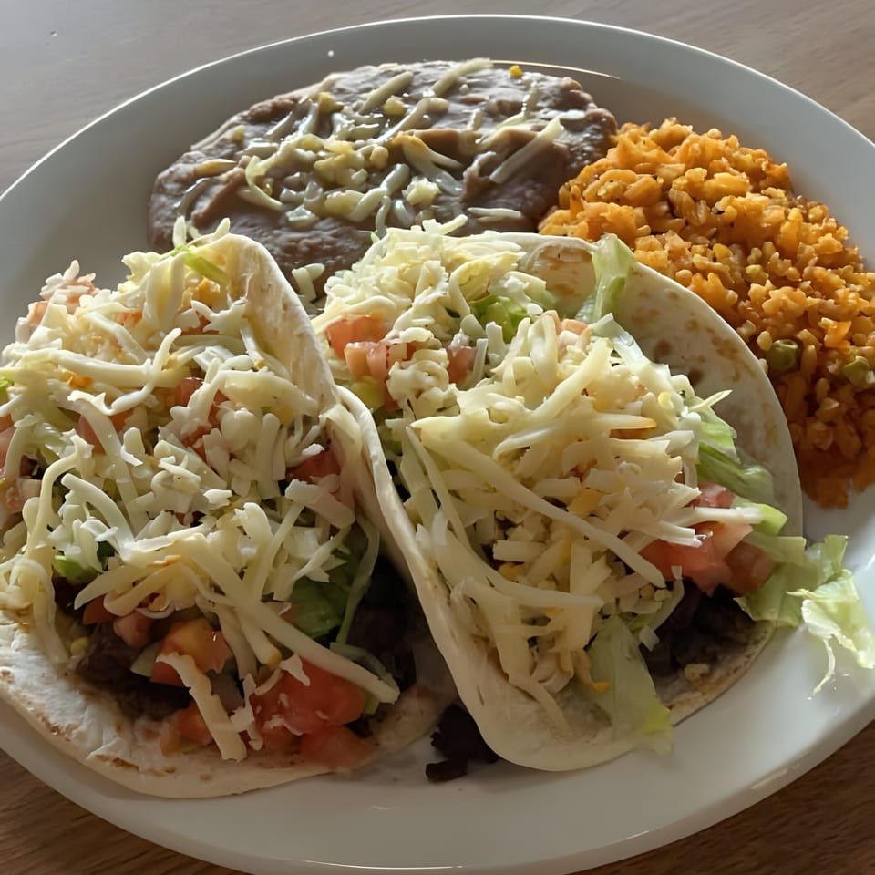 Two Mexican tacos with shredded cheese, lettuce, and tomatoes, served with rice and refried beans.